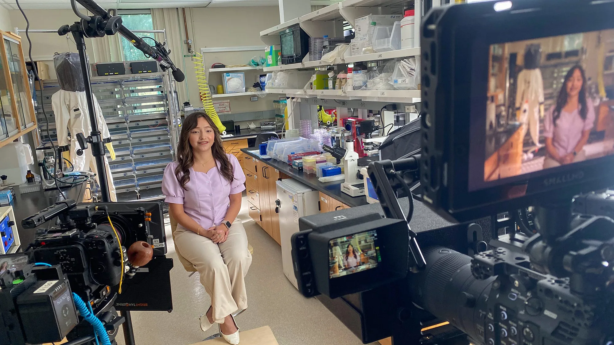 A young woman sits for an interview in a lab as the camera monitor frames her.