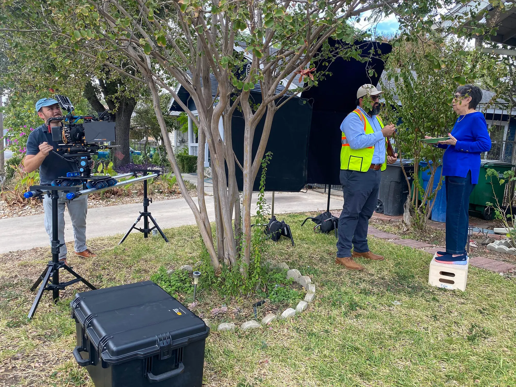 Un integrante del equipo, con chaleco de seguridad, platica con la Sra. Herrera en el jardín, frente a su casa. A la izquierda, un camarógrafo graba con una cámara montada en un slider.