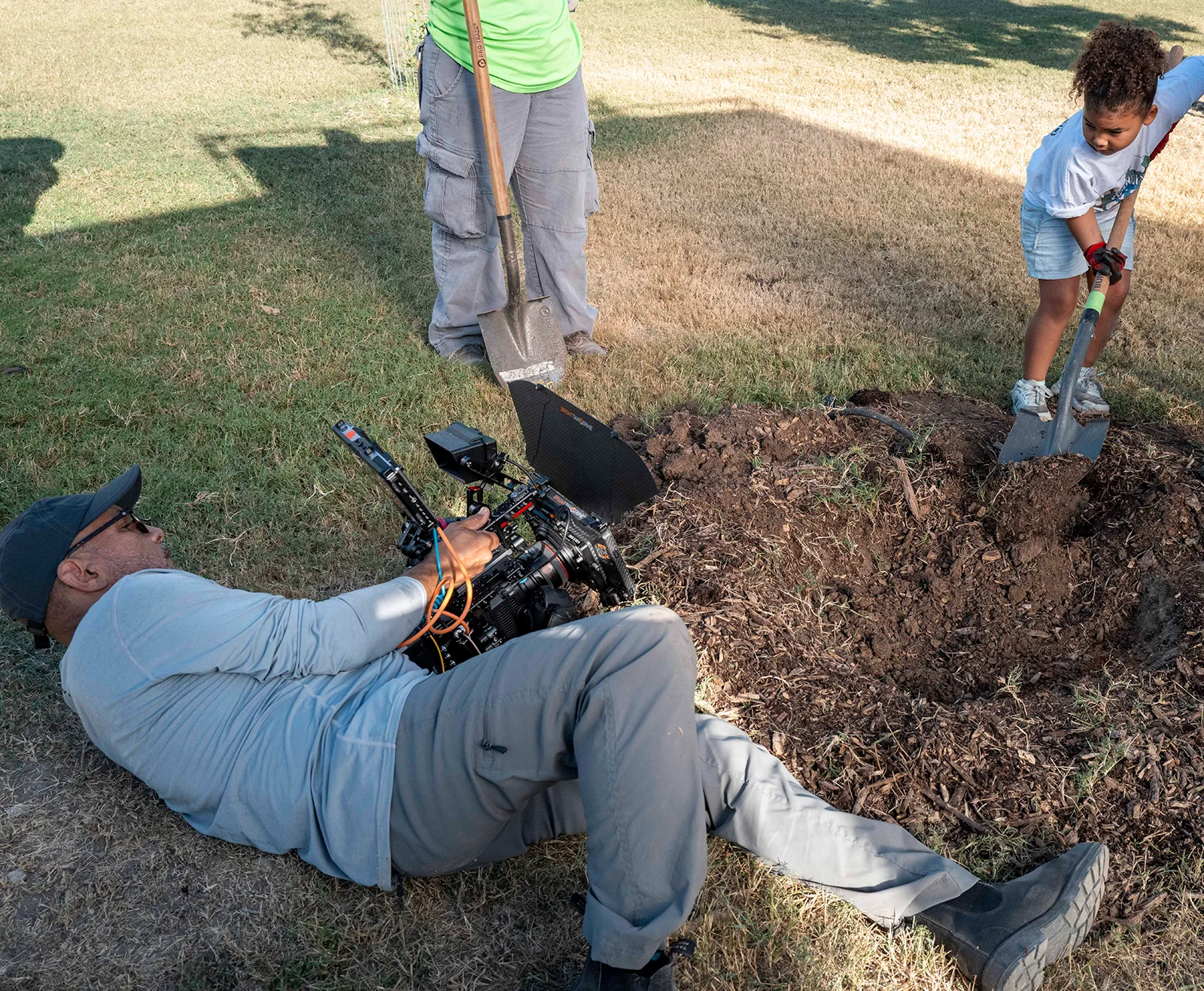 Un camarógrafo, acostado en el pasto con una cámara de cine, graba a una niña que cava un hoyo para plantar un árbol.