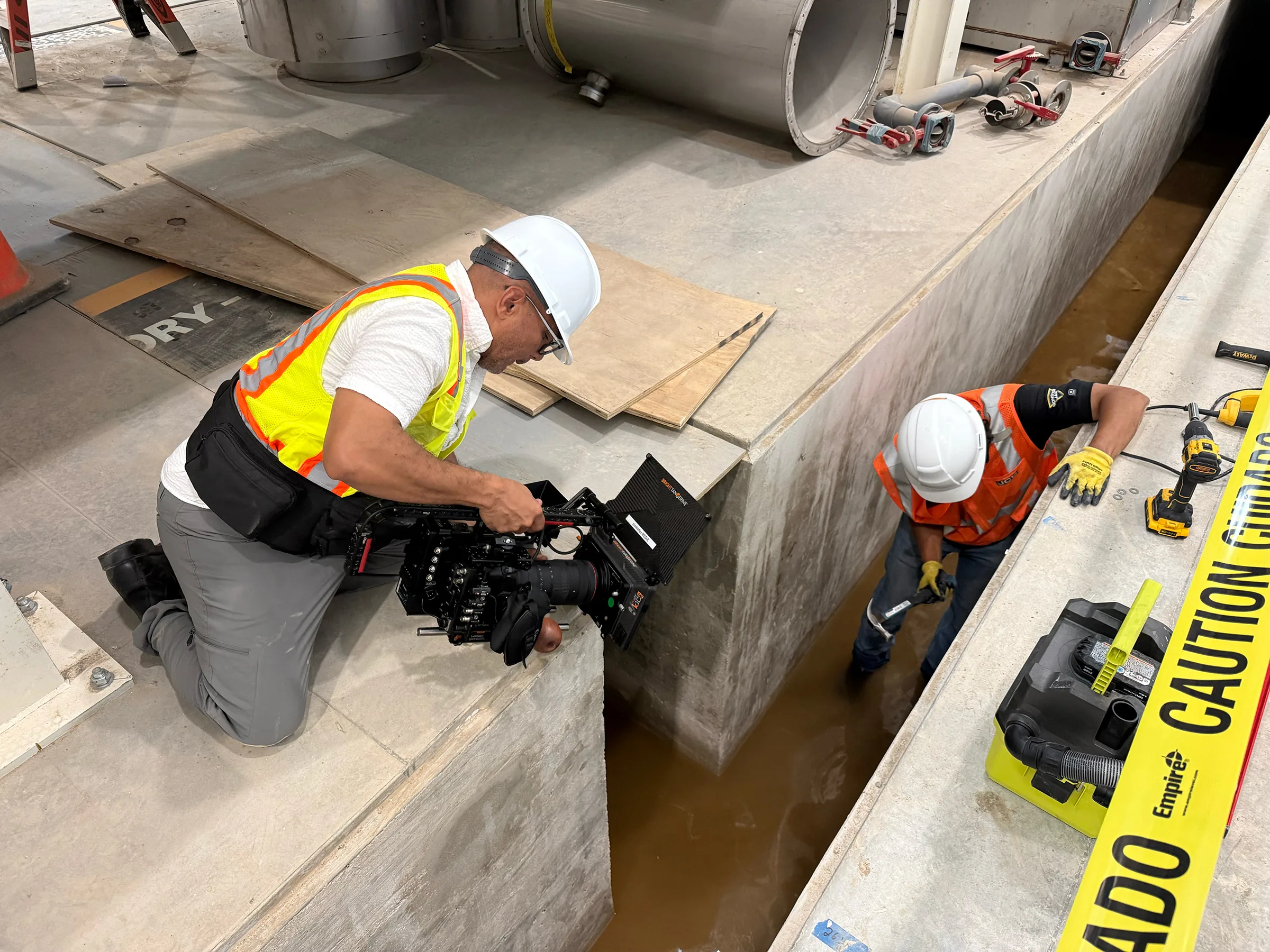 Carlos Javier Sanchez, hincado junto a una zanja de concreto, graba con una cámara de cine a un aprendiz de JCB que está abajo.