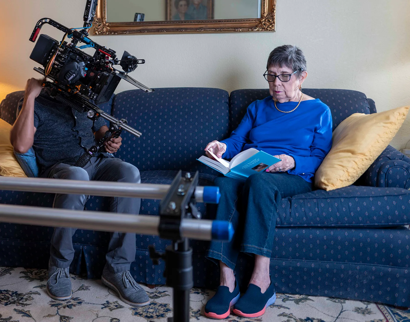 Ms. Herrera reads a book on her couch while Carlos Javier Sanchez records her with a camera on a shoulder rig in her living room.
