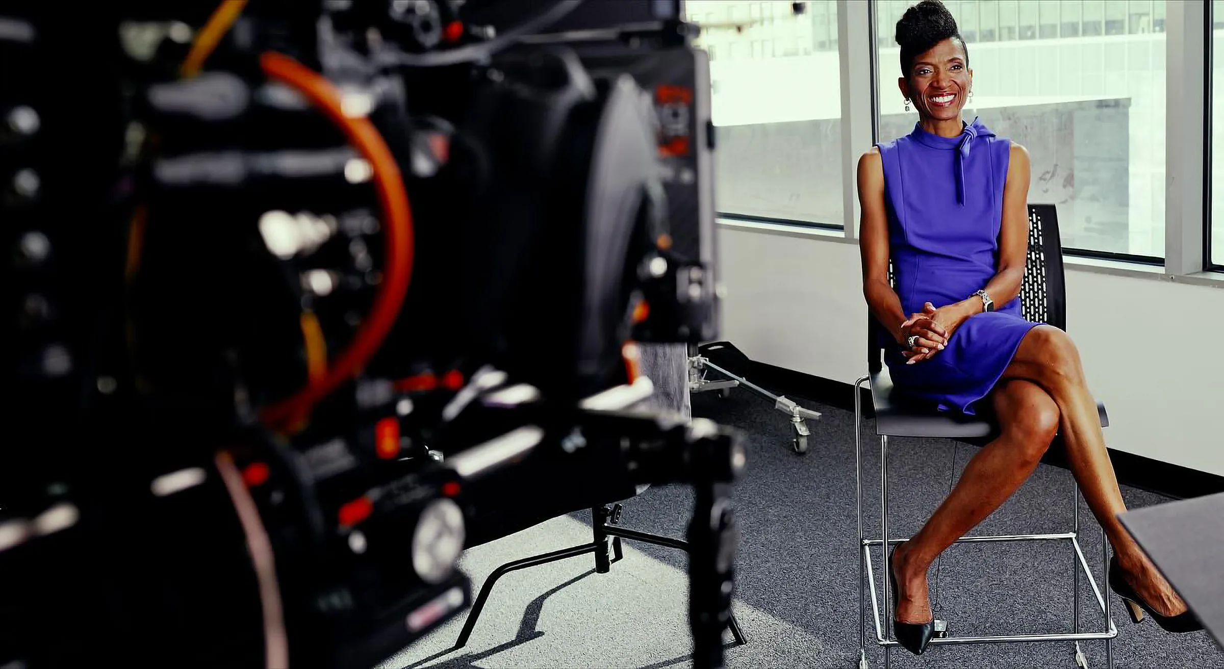 Dr. Sarah David of NPower Texas, in a purple dress, smiles from a tall chair by an office window, seen past a cinema camera.