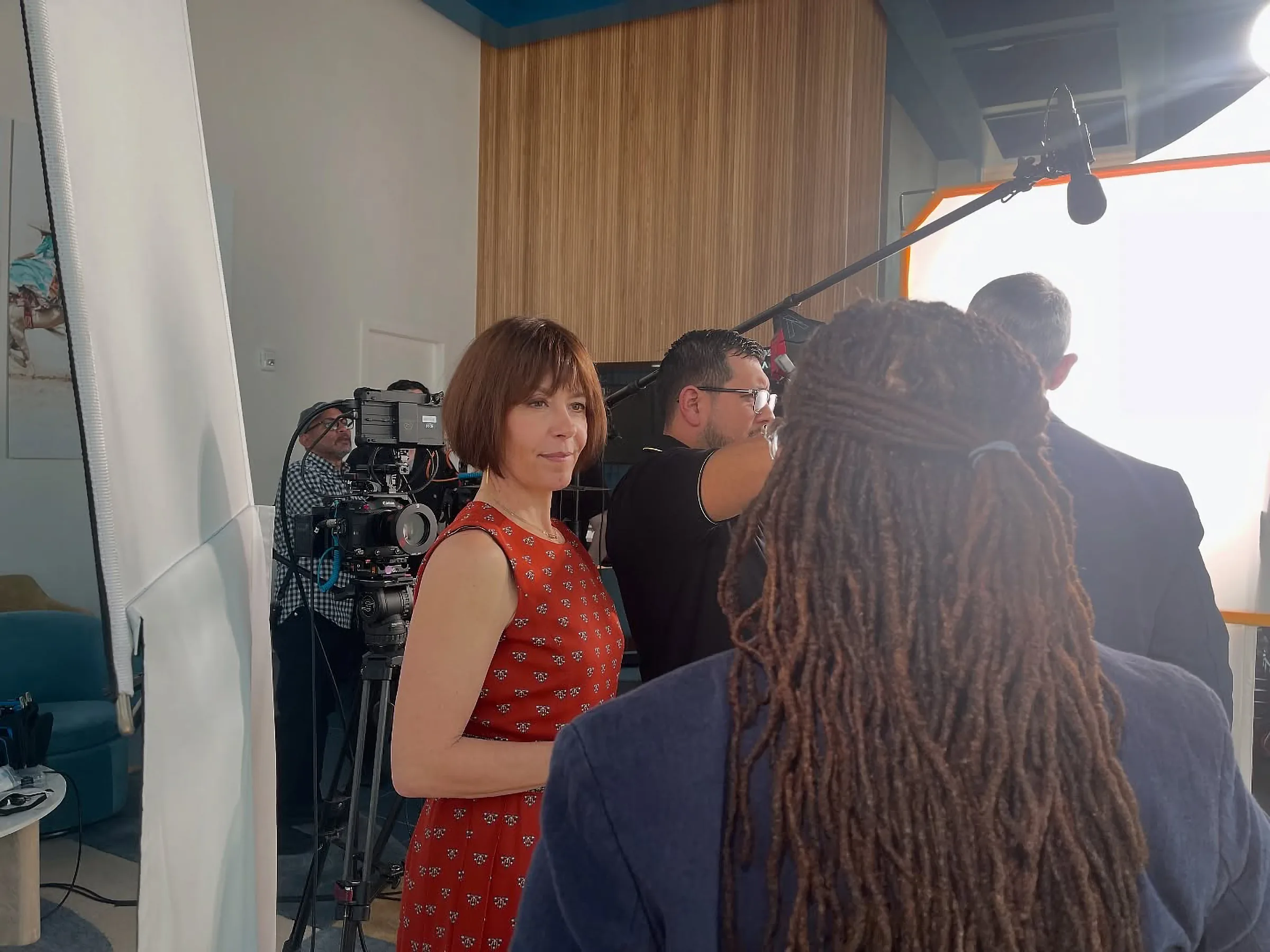 Steffi Ockenfels, de vestido rojo, platica con Sonya Bryant, que aparece de espaldas, mientras el equipo de cámara y sonido trabaja a su alrededor.