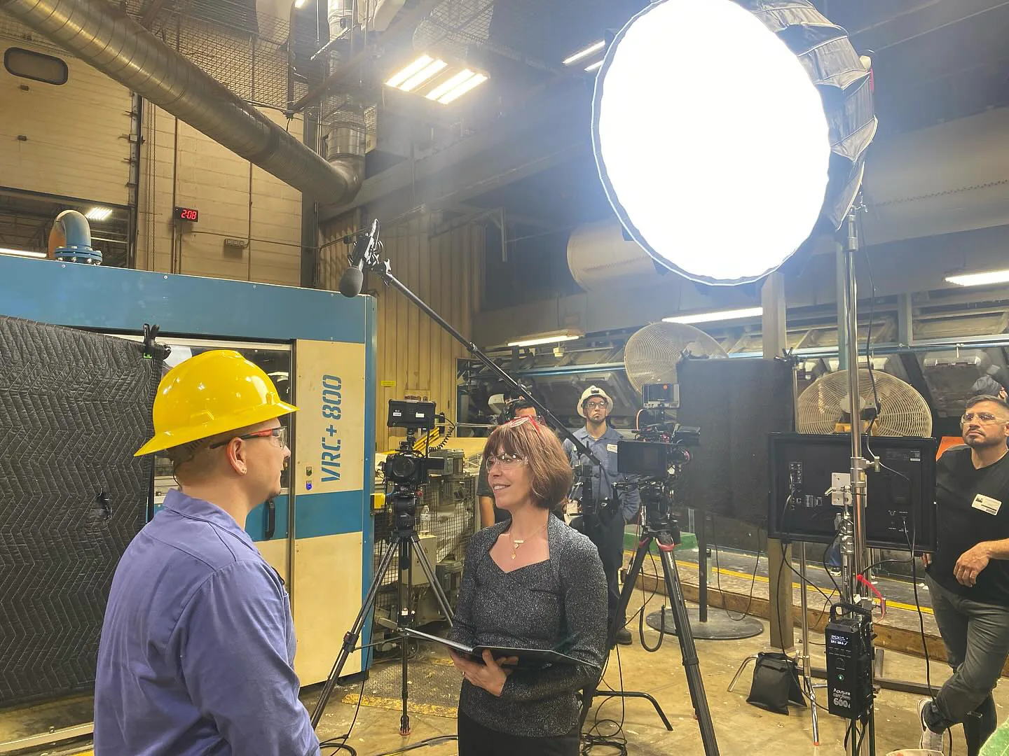 Steffi Ockenfels platica con Roman Sanchez, egresado de TX Fast Track, quien lleva casco amarillo. Los dos están bajo la luz de un softbox, en el área de producción de una fábrica.