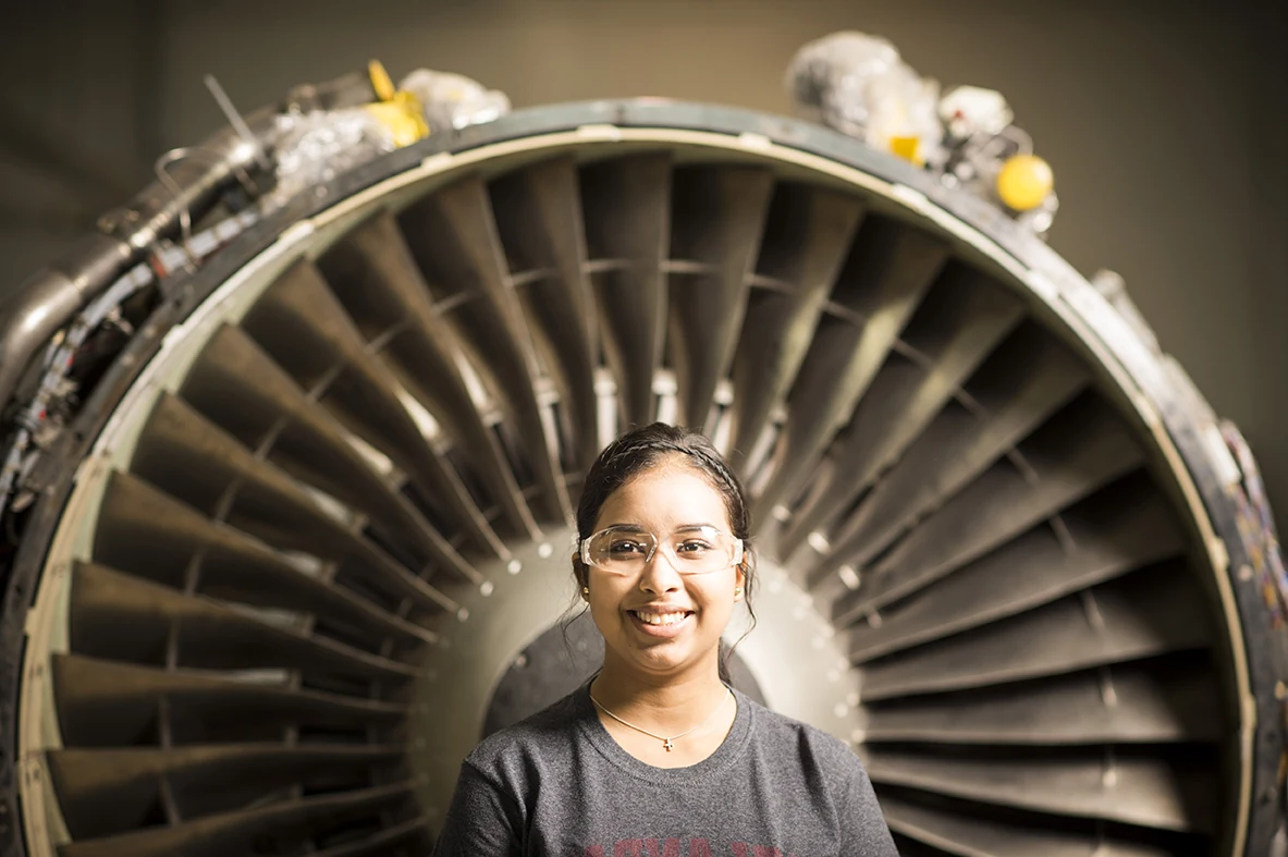 An aircraft technician student in safety glasses smiles in front of the fan blades of a jet engine at St. Philip's College.