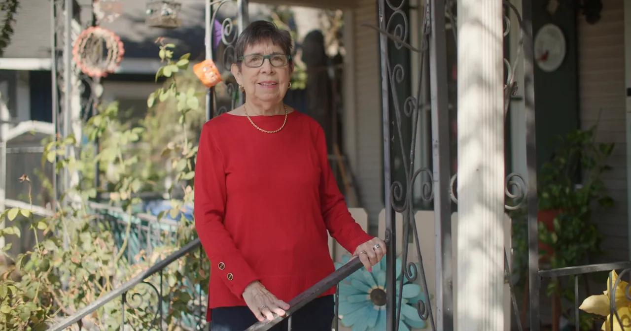 Ms. Herrera stands on the front porch of her home in a red sweater, one hand on the railing.