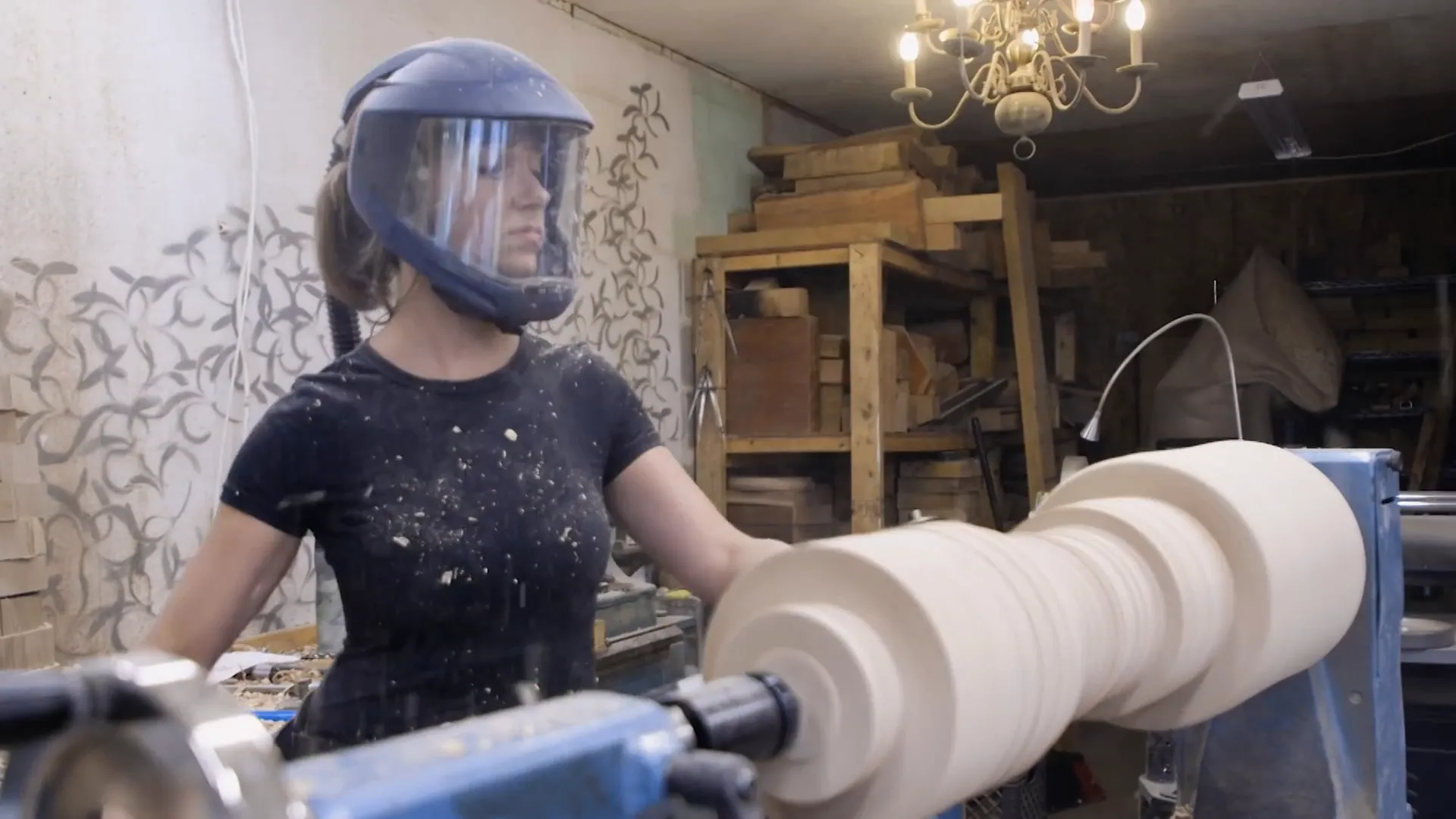 Woodturner Ashley Harwood, in a face shield, turns a large wooden piece on a lathe in her studio.
