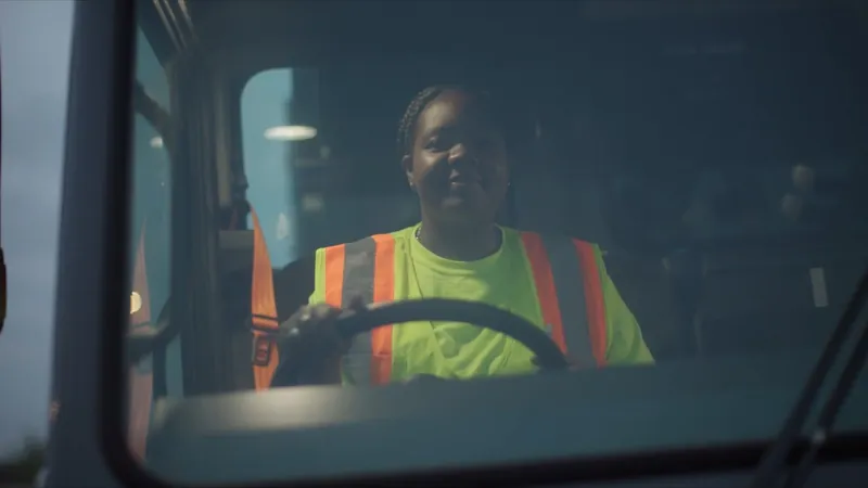 A City of San Antonio Workforce Development CDL apprentice in a safety vest smiles from behind the wheel of her truck.