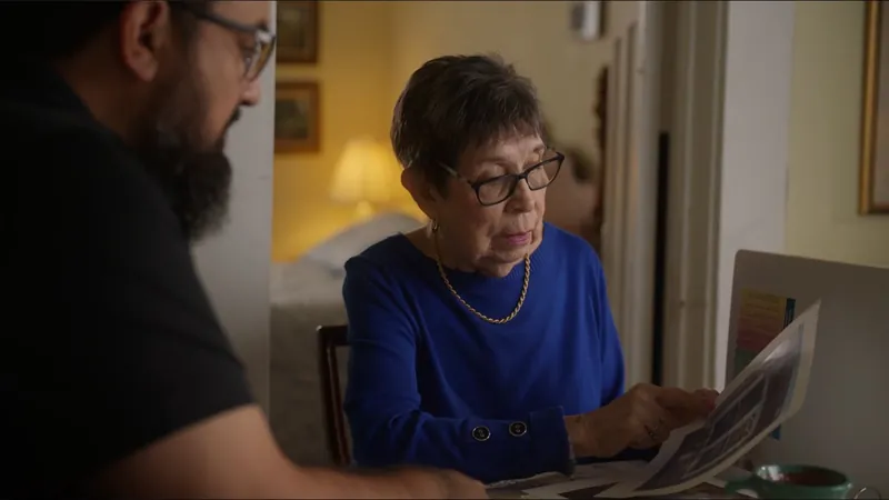 Ms. Herrera, in a blue sweater, looks over papers at her table as a man sits beside her.