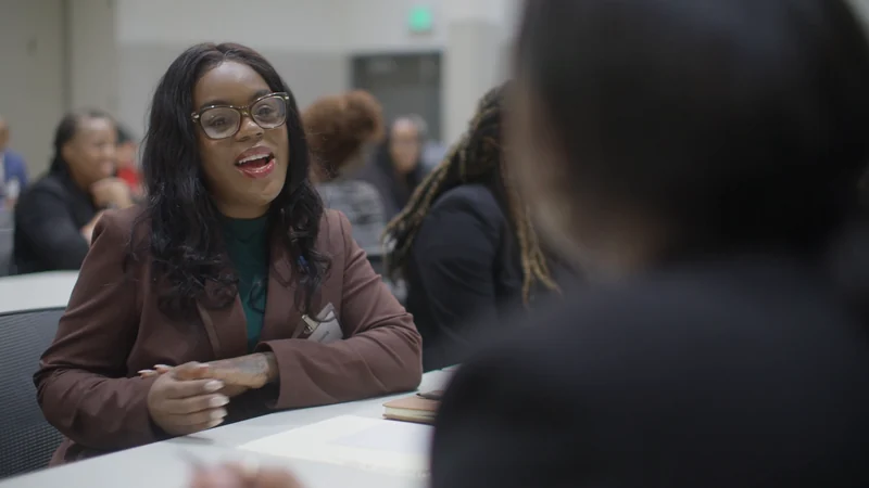 An NPower Texas student in glasses and a burgundy blazer smiles as she talks across a table, from our NPower partnership video.