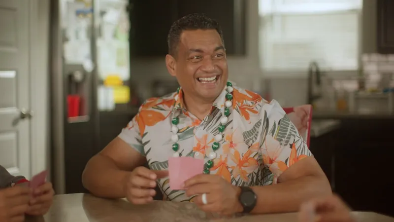 NPower alum Tas Gallahar, in a floral shirt and lei, laughs while playing cards at his kitchen table.
