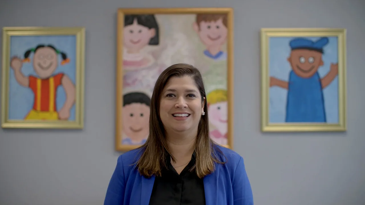 An Edgewood ISD school leader in a blue blazer smiles in front of framed children's drawings.