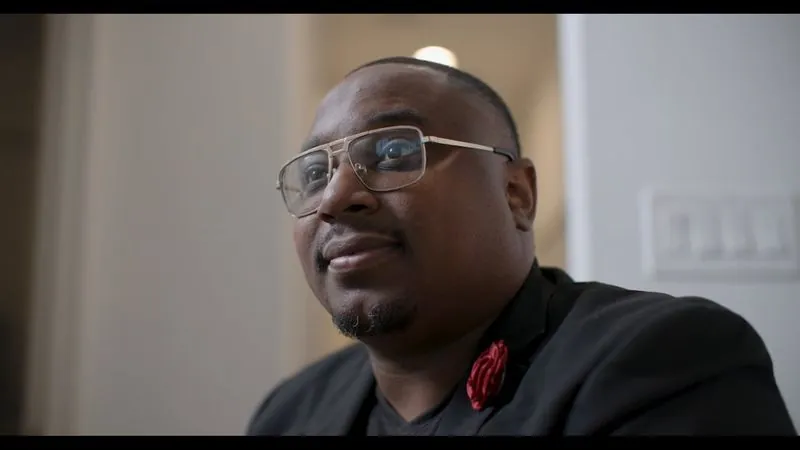 NPower alum Michael Fugett, in glasses and a dark jacket with a red flower pin, smiles during his interview.