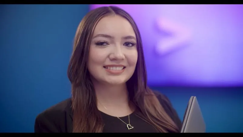 Estrella Davila, a data analyst at Accenture Federal Services and an NPower alum, smiles in front of a purple logo wall, laptop in hand.
