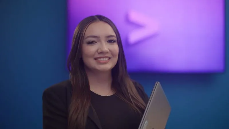 A woman from Accenture Federal Services smiles in front of a purple logo wall, laptop in hand.