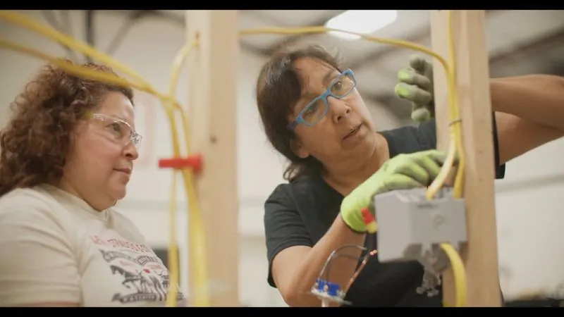 Two women wire an electrical box on a wood-frame training wall at Lone Star Construction Trades Training.