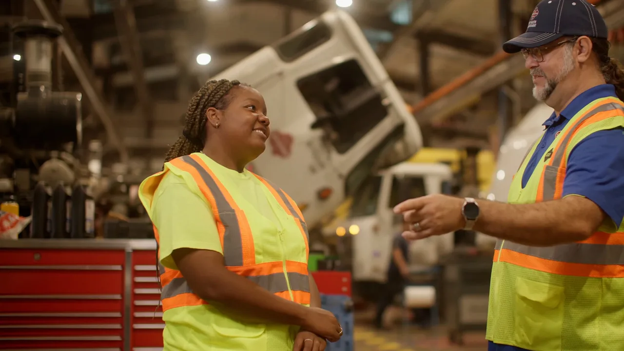 An apprentice in a yellow safety vest talks with a coworker in a truck maintenance shop.