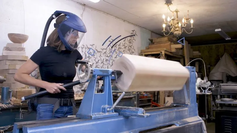 Woodturner Ashley Harwood, in a face shield, turns a large wood chandelier piece on a lathe in her studio.
