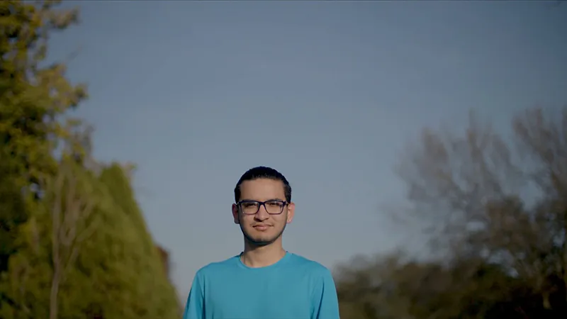 A Students of Service alum in glasses and a blue T-shirt stands outdoors against an evening sky.