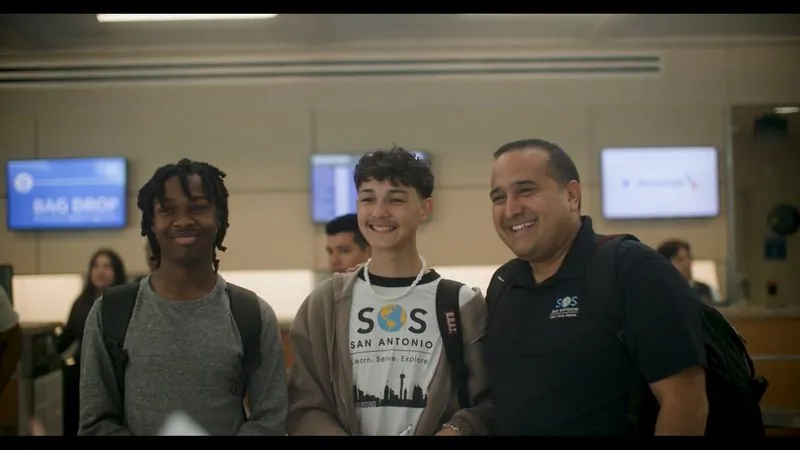 Two students, one in an SOS San Antonio shirt, smile with a program leader at an airport check-in counter.
