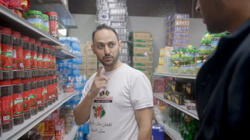 A grocer in an Afghan Village T-shirt talks with a visitor between shelves of imported goods.