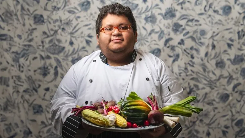 Jacob, in a white chef’s coat, holds a platter of fresh vegetables against floral wallpaper.