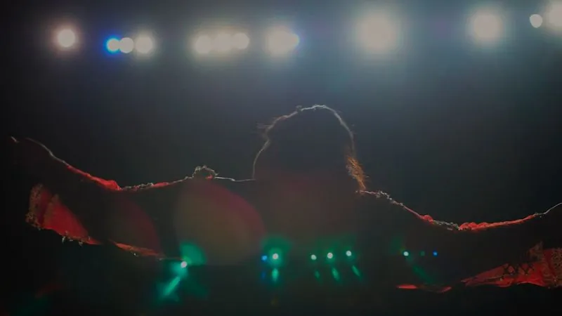 A performer, seen from behind, spreads her arms toward the stage lights in a dark theater.