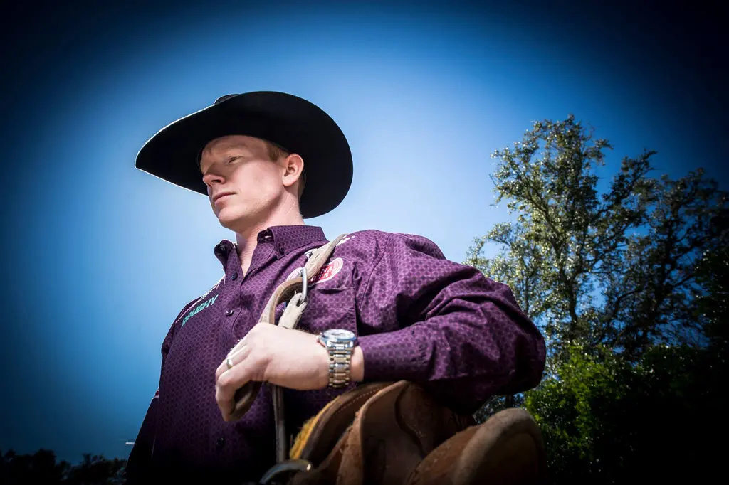 Rodeo cowboy in a black hat and purple western shirt holding his saddle against a blue sky