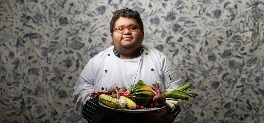 Jacob Gutierrez, in a white chef’s coat, holds a platter of fresh vegetables against floral wallpaper.