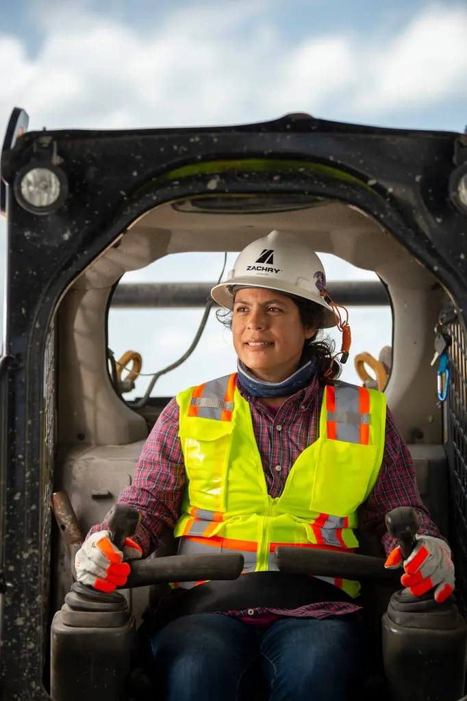 Evelyn Sanchez, a Zachry Construction equipment operator, in a white hard hat and yellow safety vest at the controls of a loader