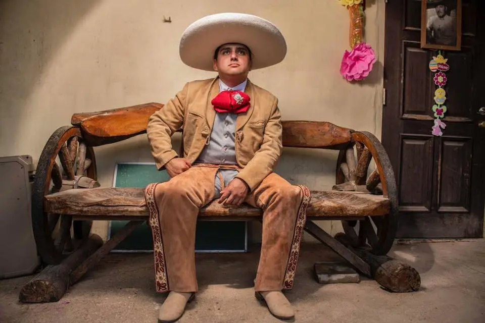 Young charro in a tan suede suit, red bow tie and white sombrero seated on a wagon-wheel bench
