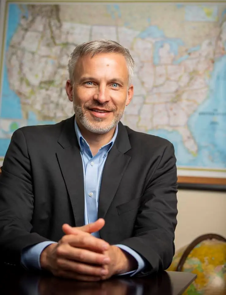 Mike Larson in a dark blazer smiling at a desk in front of a wall map of the United States