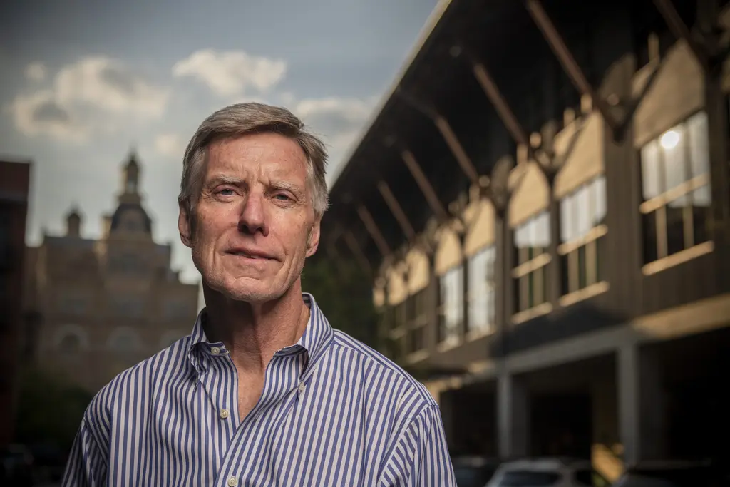 Frank Burney in a blue striped shirt stands outdoors with a historic tower and a timber-framed building behind him