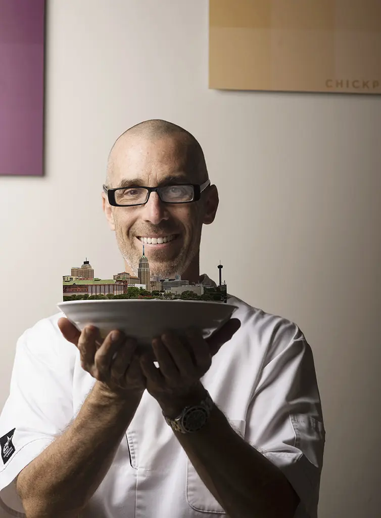 Andrew Weissman in glasses and chef's whites holds a small architectural model on a plate
