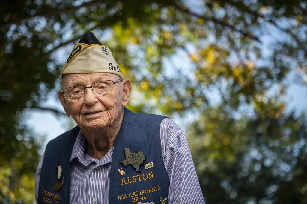 J.C. Alston, an elderly Navy veteran in glasses, a veterans cap and USS California vest, outdoors under trees