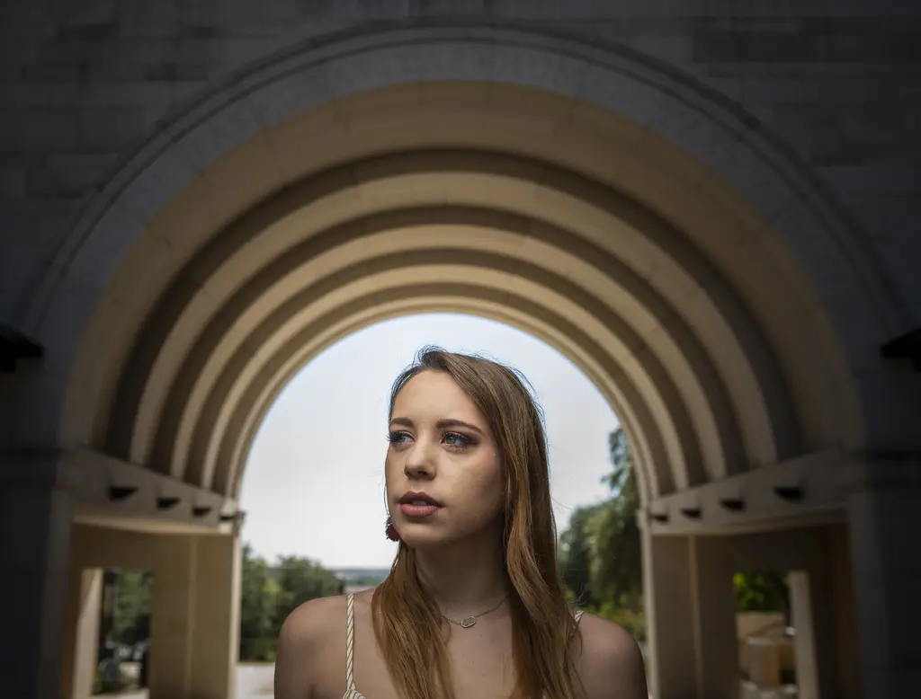 Young woman with long hair standing beneath a layered stone archway against an overcast sky