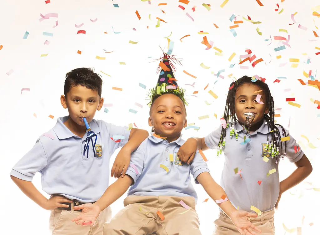 Three children in light blue school uniforms, one in a party hat, celebrate as confetti falls on a white backdrop