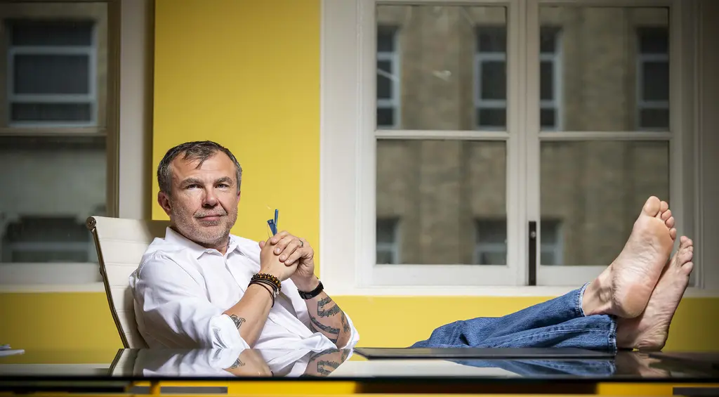 Chris Hotchkiss in a white shirt and jeans leans back with his bare feet on a table against a yellow wall