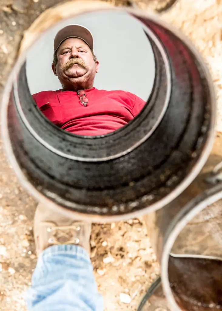 Bill Seger in a red shirt and cap reflected in crude oil inside a bucket, his boot on the ground below