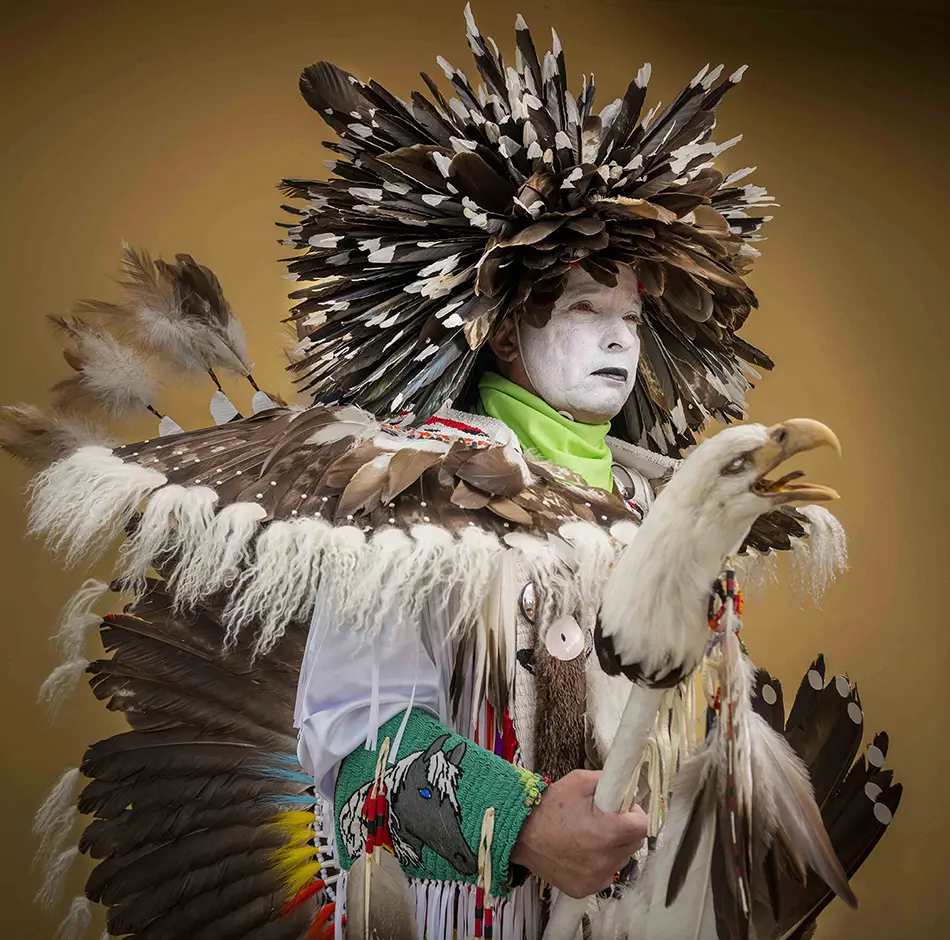 Dancer in white face paint and feathered eagle regalia posing against a warm brown backdrop