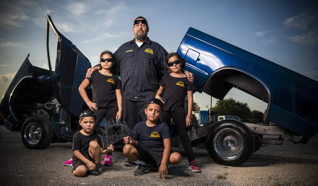 Man and four children in black shirts posing before a blue lowrider truck with its doors and bed raised