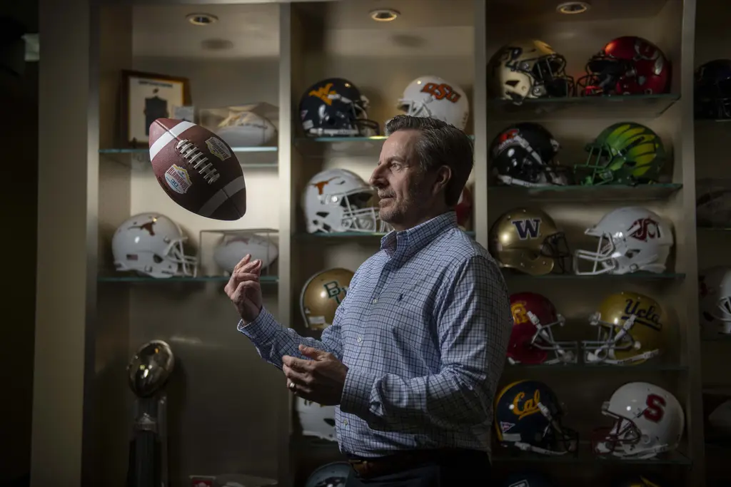 Derrick Fox tossing a football in front of shelves of college football helmets