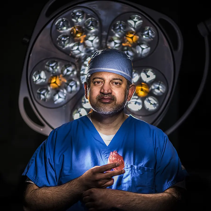 Dr. Muhammad A. Mumtaz in blue scrubs and a surgical cap holds a model heart beneath operating room lights