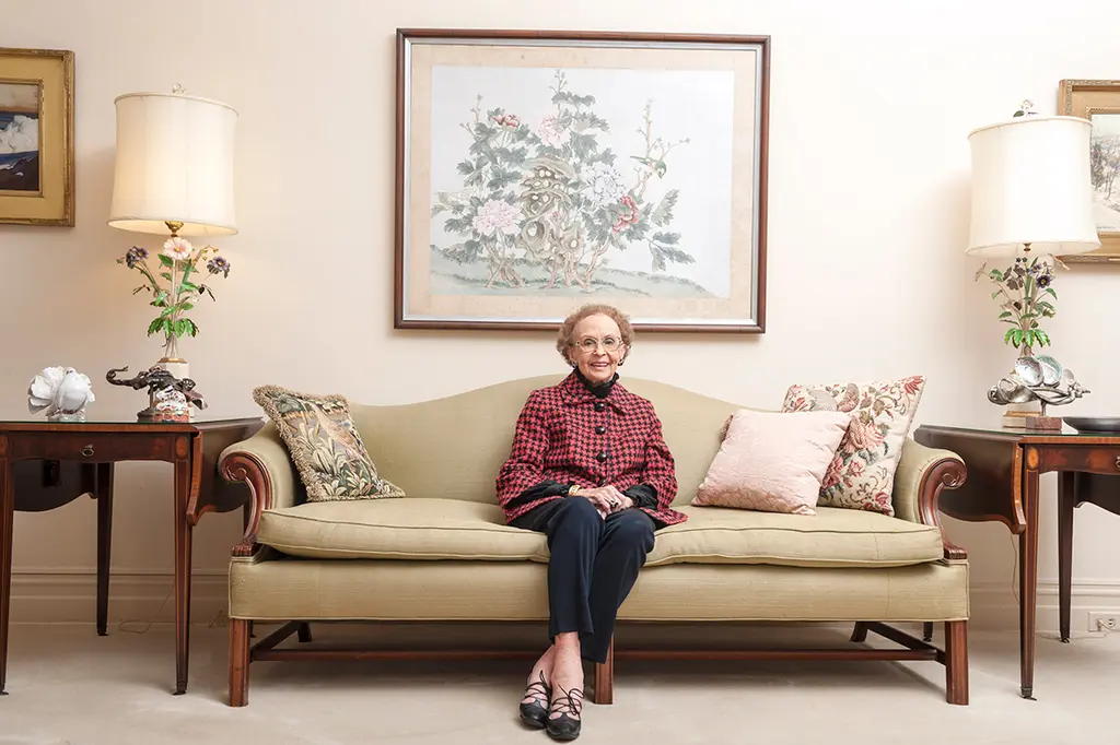 Edith McAllister in a red plaid shirt sits alone on a camelback sofa between two lamps in a formal living room