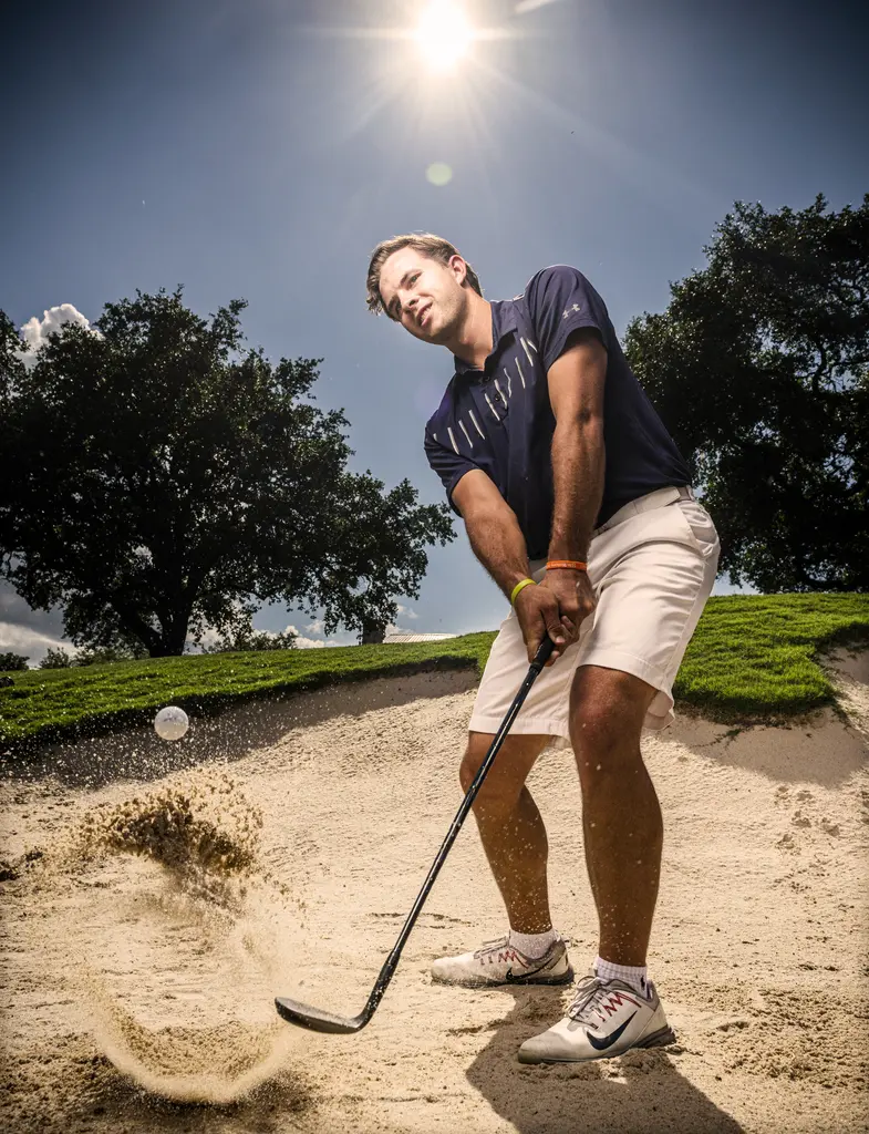 Garrett Martin in a navy shirt and white shorts swings through a bunker shot as sand sprays under a bright sun