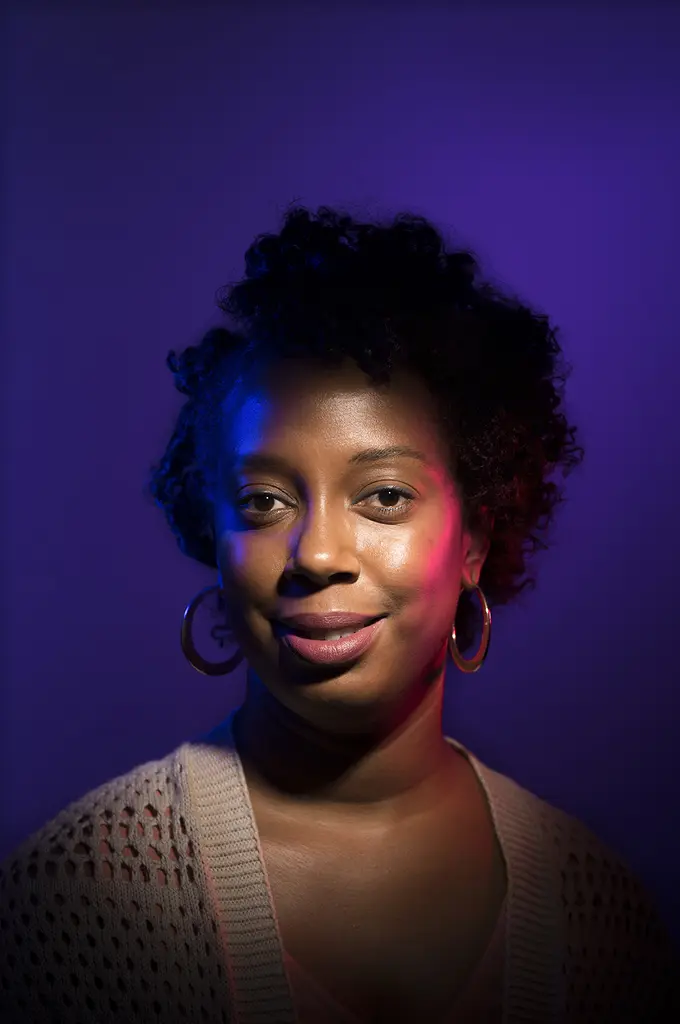 Kristen Cates, with short curly hair and gold hoop earrings, lit by purple, blue and red light on a violet backdrop