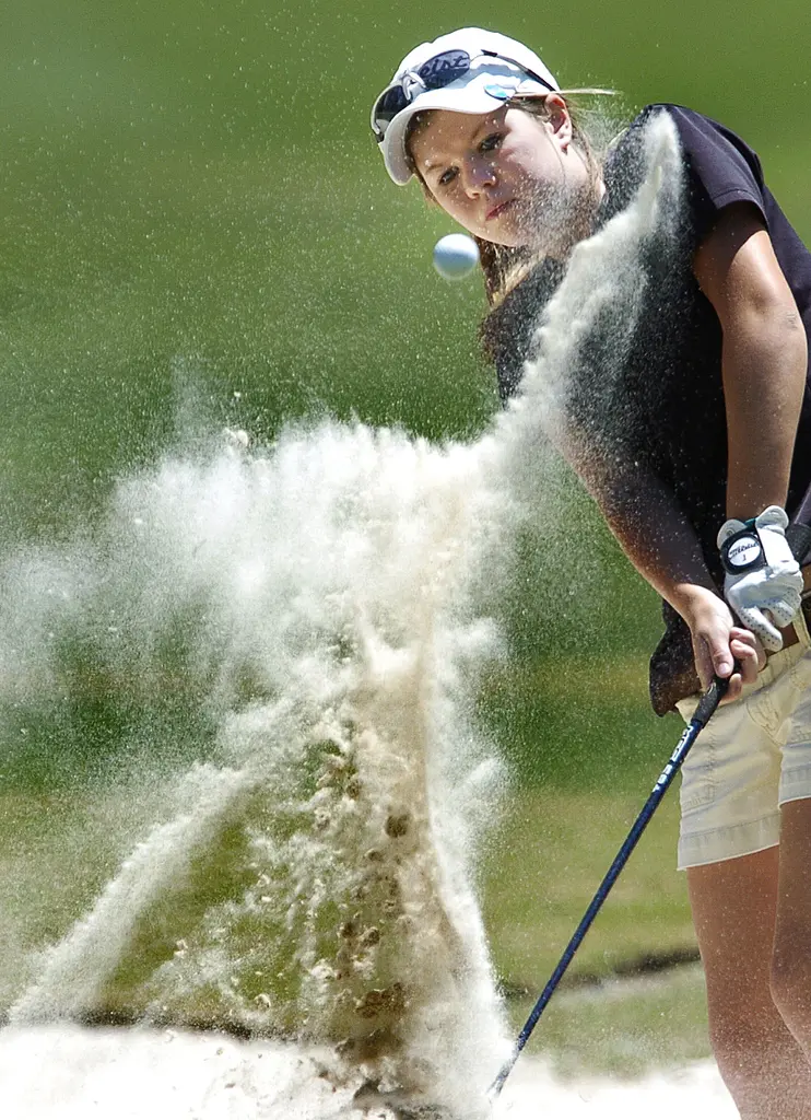 Dina O'Brien, in a white visor, blasting a golf ball out of a bunker in a spray of sand