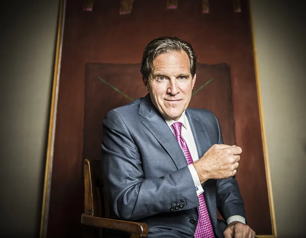Jeff Garvens in a gray suit and pink tie seated in a tall high-backed wooden chair, one fist at his chest