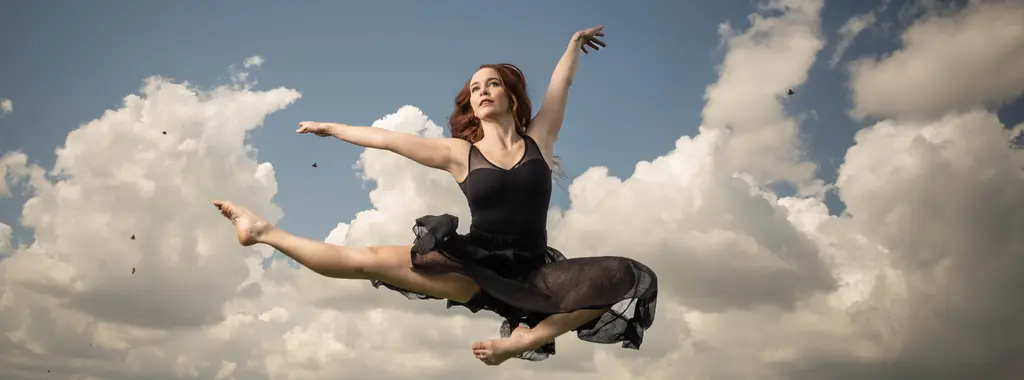 Dancer Jenna Mathews in a black dress leaps into a midair split against a blue sky with white clouds
