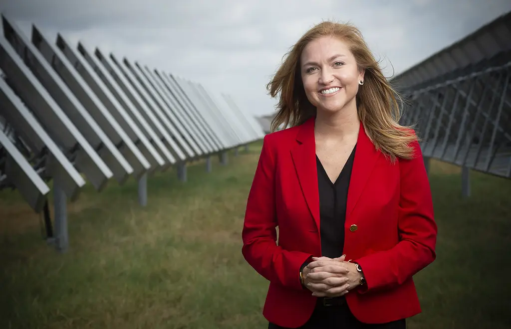 Jenna Saucedo-Herrera in a red blazer smiles between rows of solar panels under a cloudy sky